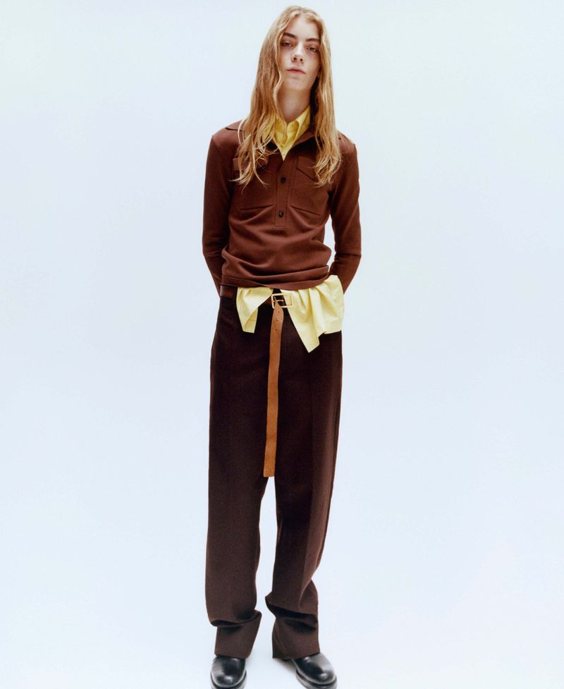 Model with long blonde hair wearing a brown long-sleeve shirt over a yellow shirt and brown trousers against a white seamless backdrop.