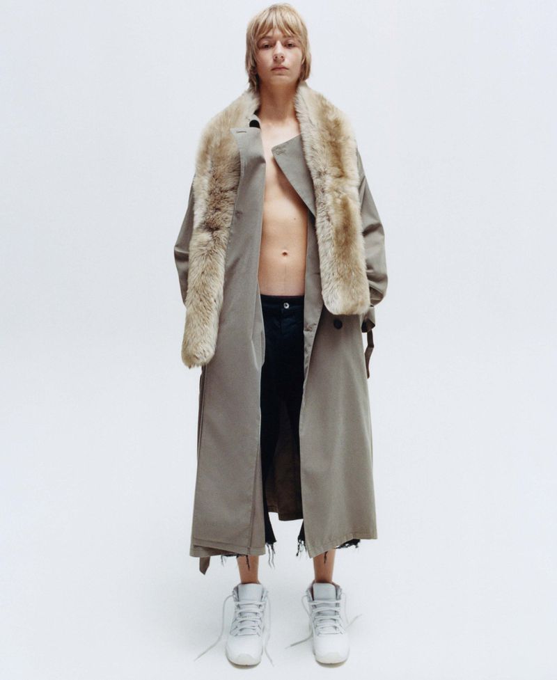 Model wearing a long taupe coat with a fur stole and white sneakers against a plain white studio backdrop.