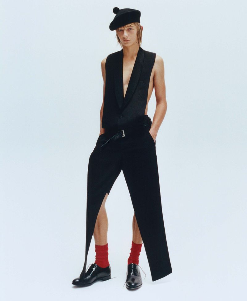 Model wearing a black beret, sleeveless black vest, trousers, red socks, and black shoes against a white background