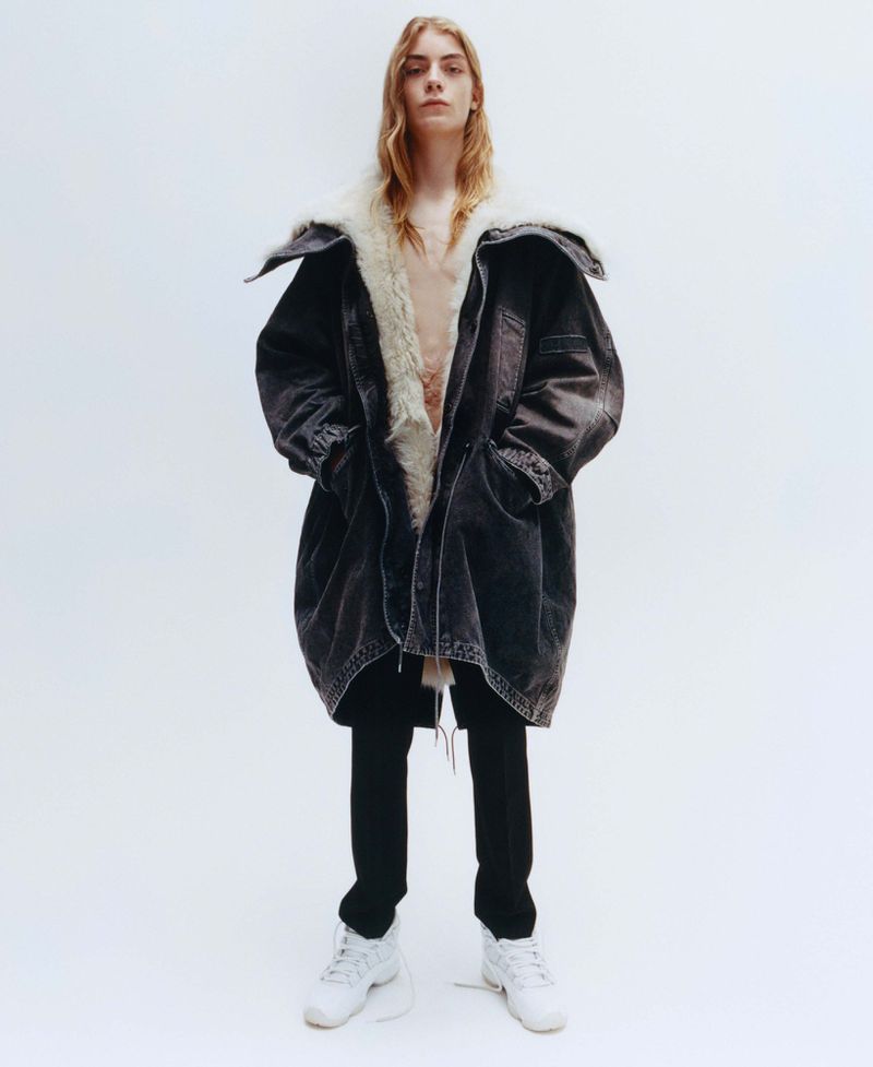 Model with long blonde hair wearing a dark shearling-lined coat and black trousers against a plain white background.