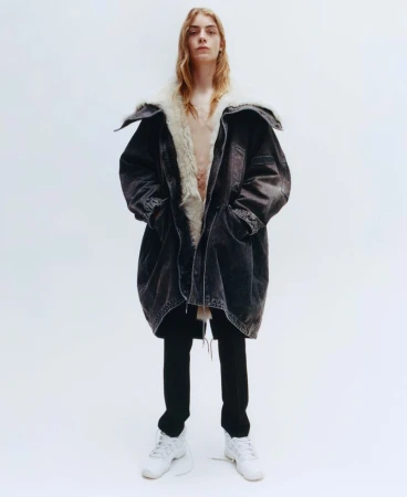 Model with long blonde hair wearing a dark shearling-lined coat and black trousers against a plain white background.