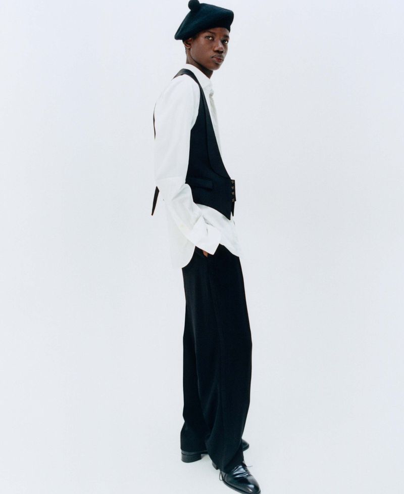 Model wearing a black beret, white shirt, dark vest, and trousers against a clean white seamless studio backdrop.