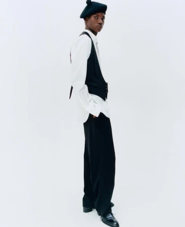 Model wearing a black beret, white shirt, dark vest, and trousers against a clean white seamless studio backdrop.