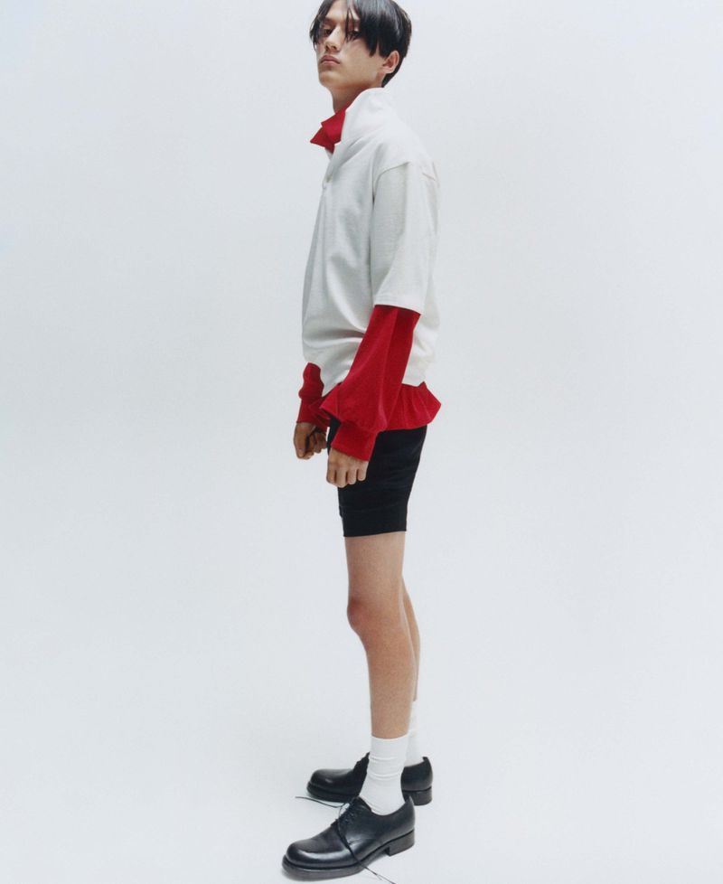 Young male model posing in profile wearing a layered white and red shirt against a plain white background.