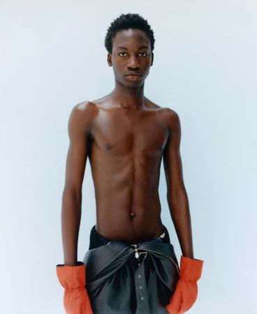 Shirtless black male model wearing dark trousers and orange gloves against a light background for M Le magazine du Monde.
