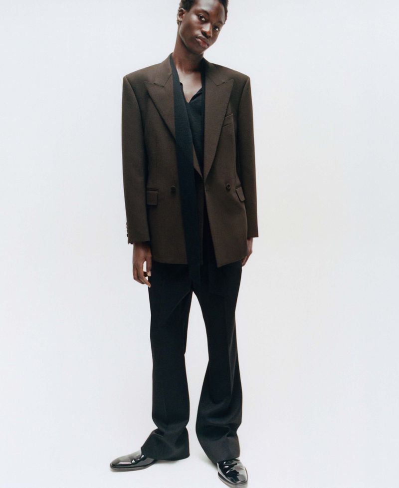 Model wearing a brown blazer and black trousers against a neutral white studio background for an editorial spread.