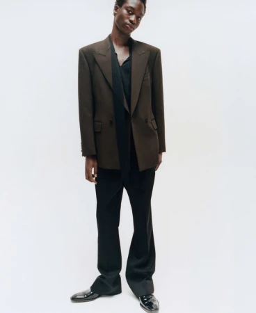 Model wearing a brown blazer and black trousers against a neutral white studio background for an editorial spread.