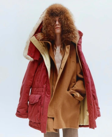Model wearing a layered red parka with a fur hood and a brown coat against a white background