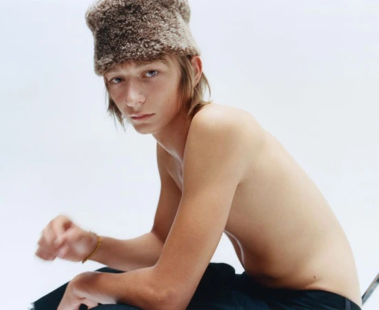 Shirtless male model wearing a furry brown winter hat against a plain white studio background