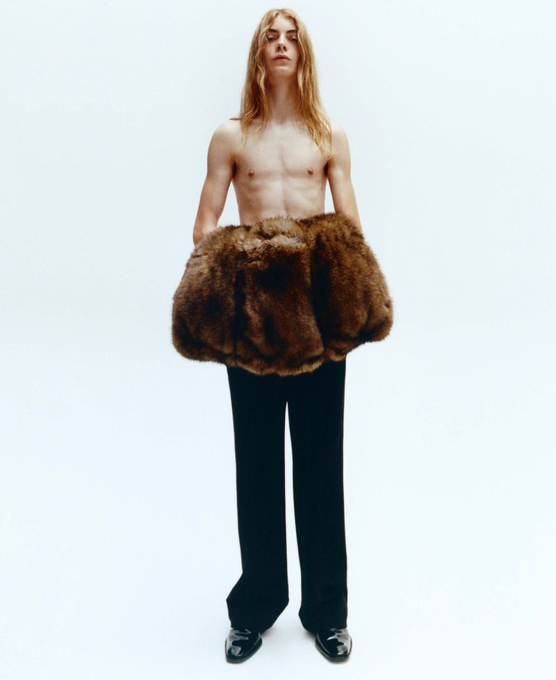 Model with long blonde hair standing shirtless wearing a large brown fur muff and black trousers against a plain white background
