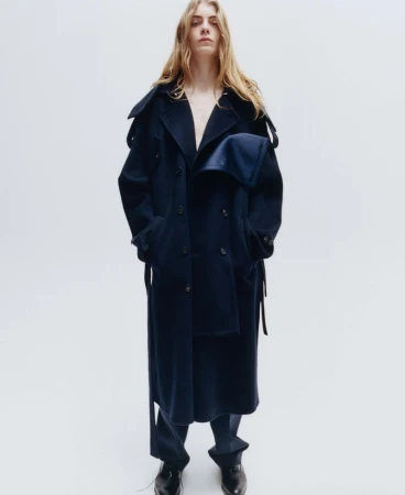 Model with long blonde hair wearing a dark navy wool coat against a plain white studio background.