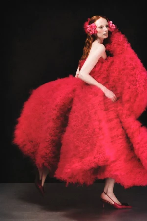 Model in voluminous red ruffled dress with matching floral hair accessories against a dark background