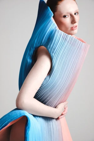Model wearing a structured pleated blue and pink garment against a plain white background