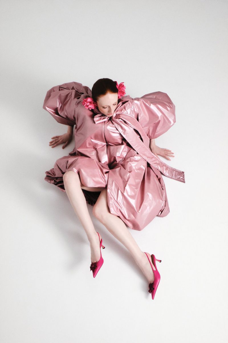 Model wearing a glossy pink puff-sleeve dress and pink heels seated against a plain white background
