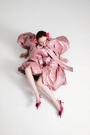 Model wearing a glossy pink puff-sleeve dress and pink heels seated against a plain white background