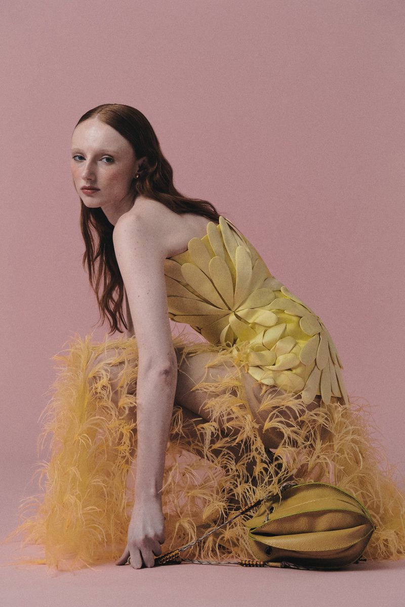 Model in a sculptural yellow petal dress with feathered hem posing against a solid dusty pink studio backdrop.