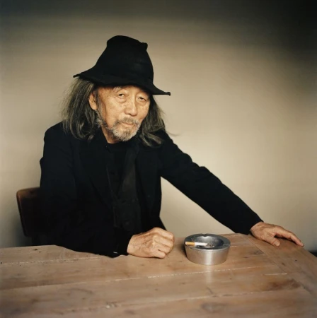 Portrait of Yohji Yamamoto in a black hat and jacket seated at a wooden table with an ashtray