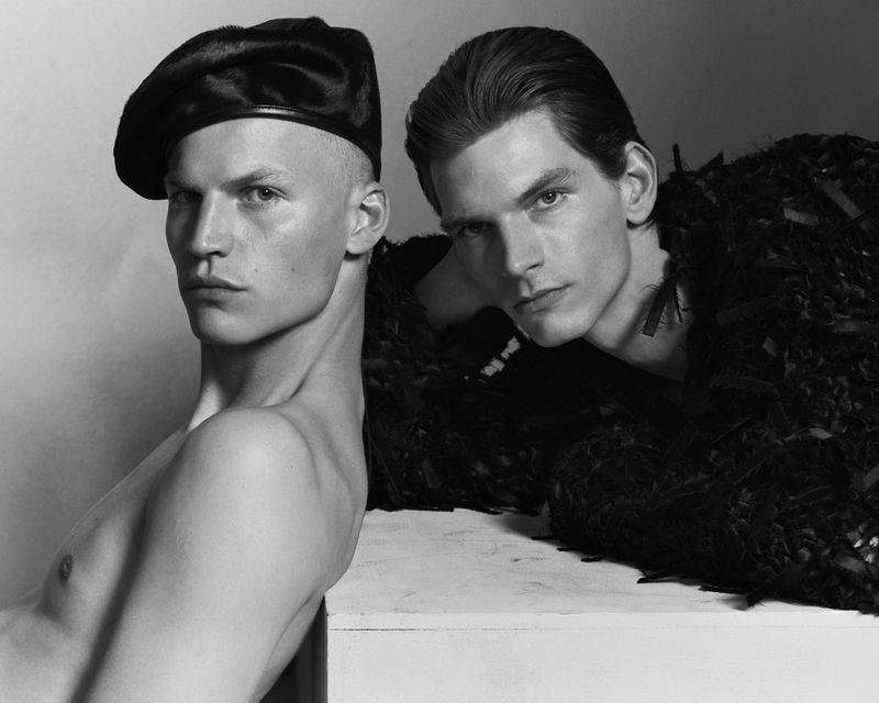 Black-and-white studio portrait of two slender adult male fashion models posing with serious expressions, one wearing a beret and textured garment.