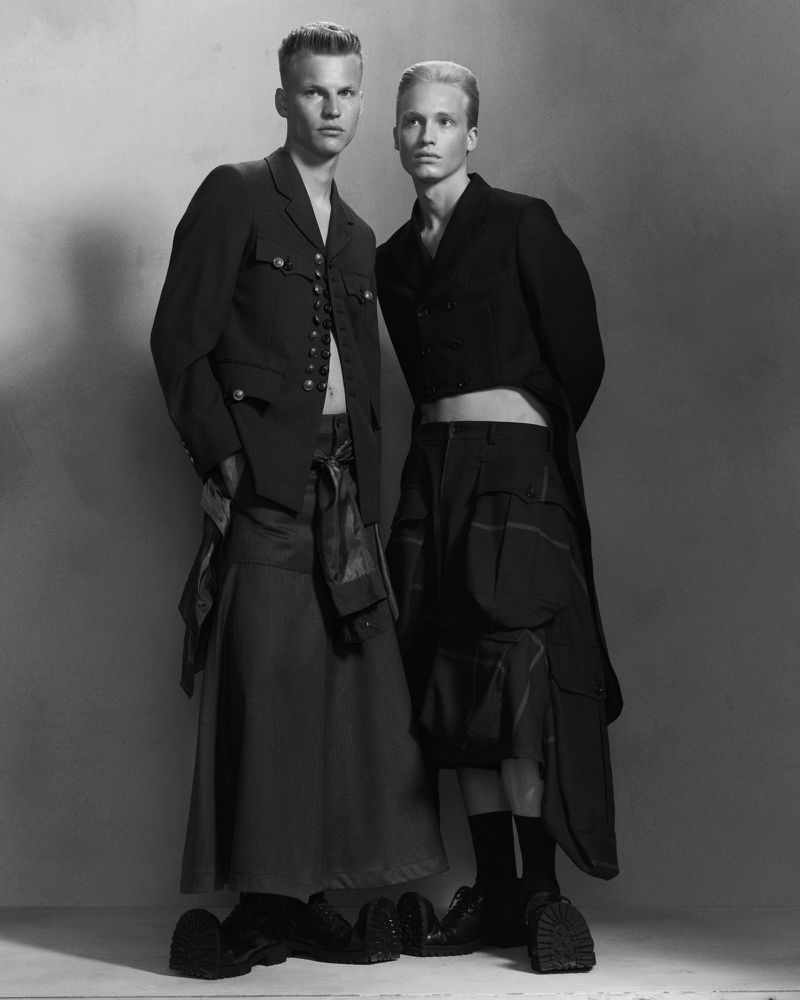 Two young adult male models in formal jackets and skirts pose in a black-and-white fashion studio shot, showcasing androgynous clothing.