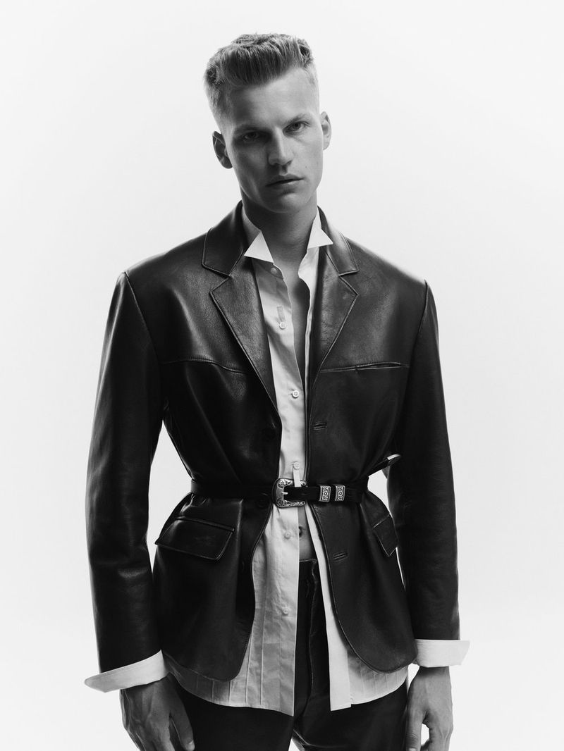 A black and white portrait of a young adult male wearing a black leather blazer cinched with a black western-style belt against a white backdrop.