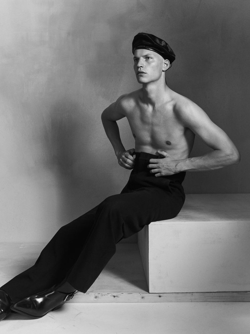 Shirtless male model wearing a black beret and high-waisted trousers seated on a white block in a studio.