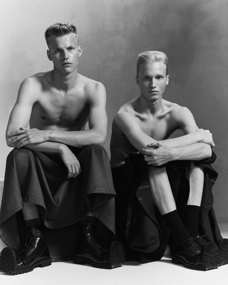 Two shirtless male models seated in black draped skirts and lug-sole shoes against a grey studio backdrop.