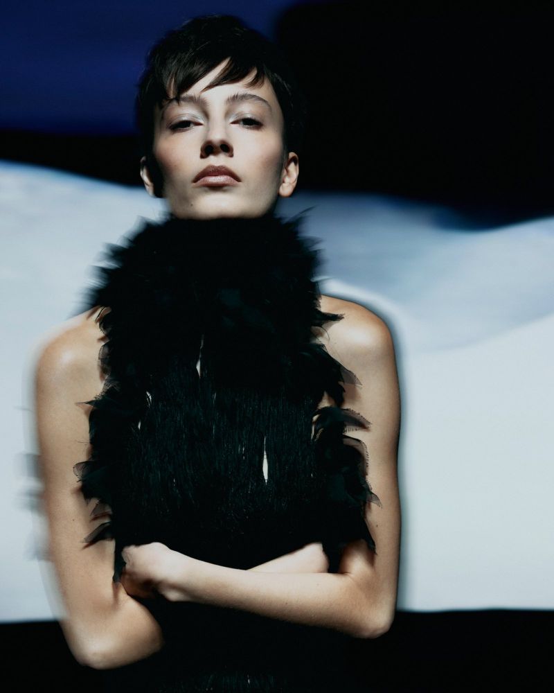 Model with short dark hair wearing a black feathered top against a dark and blurred background for Vogue Portugal.