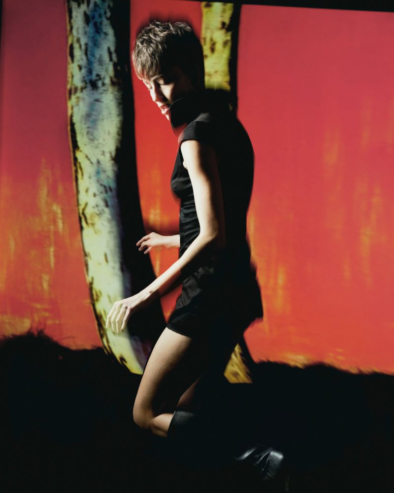 Model in a black dress posing against a vibrant red backdrop with abstract vertical pillars for Vogue Portugal.