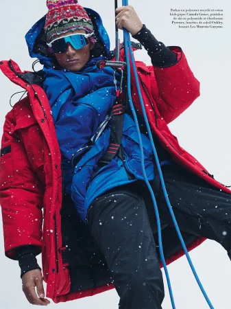 Male model in red jacket, blue parka, and patterned beanie wearing mirrored sunglasses while holding climbing ropes in falling snow.