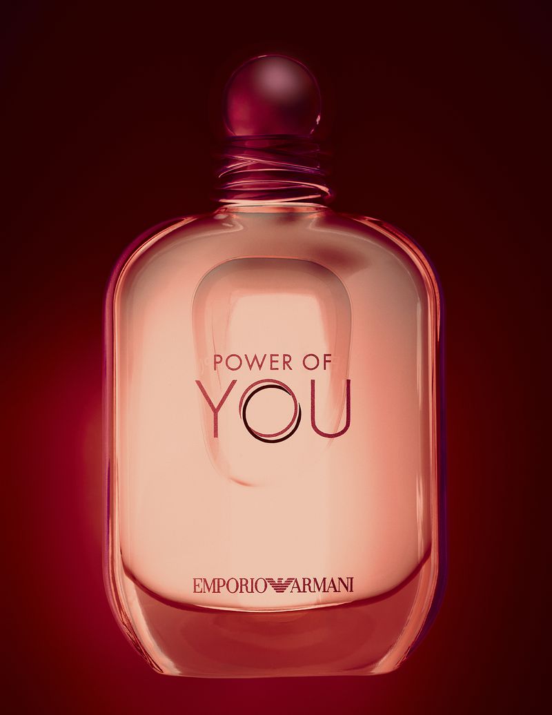 Close-up product photograph of a transparent pink glass perfume bottle against a dark red gradient background.