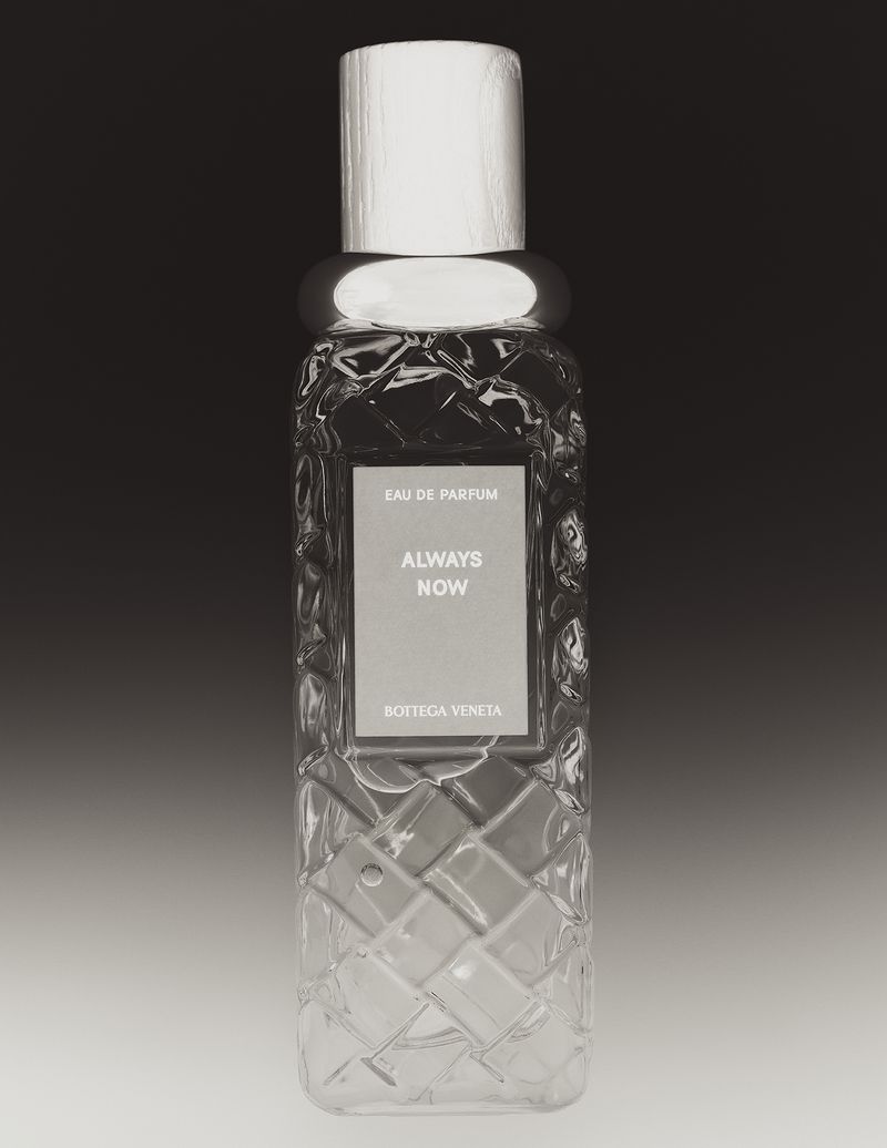 Close-up product photograph of a clear glass perfume bottle with an intricate woven pattern and light wooden cap against a dark gradient background.