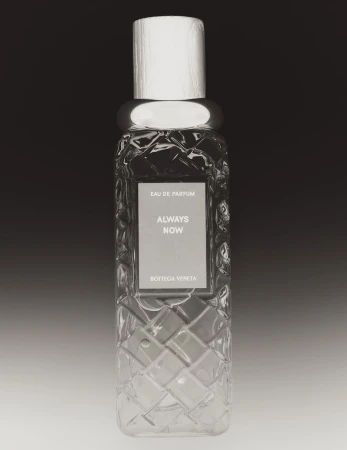 Close-up product photograph of a clear glass perfume bottle with an intricate woven pattern and light wooden cap against a dark gradient background.