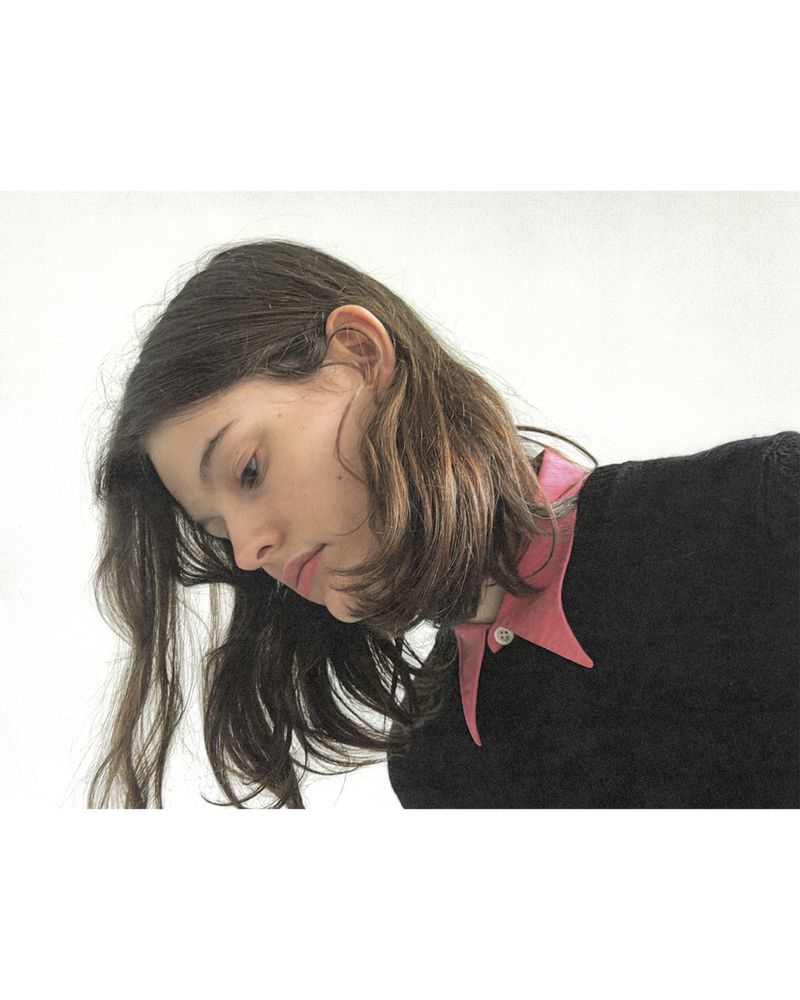 Model with long dark hair wearing a black sweater with a pink collared shirt against a white backdrop.