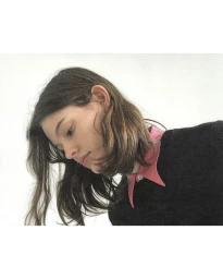 Model with long dark hair wearing a black sweater with a pink collared shirt against a white backdrop.