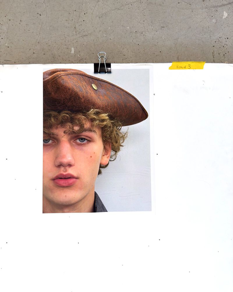 Close-up portrait of a curly-haired model wearing a brown leather tricorn hat clipped to a white backing board
