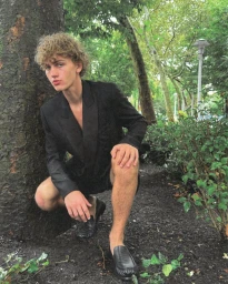 Model with curly blonde hair crouches by a tree wearing a black blazer and dark loafers in a park setting.