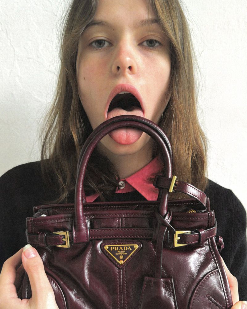 Model holding a glossy burgundy Prada handbag up to her mouth with tongue out for Numéro New York