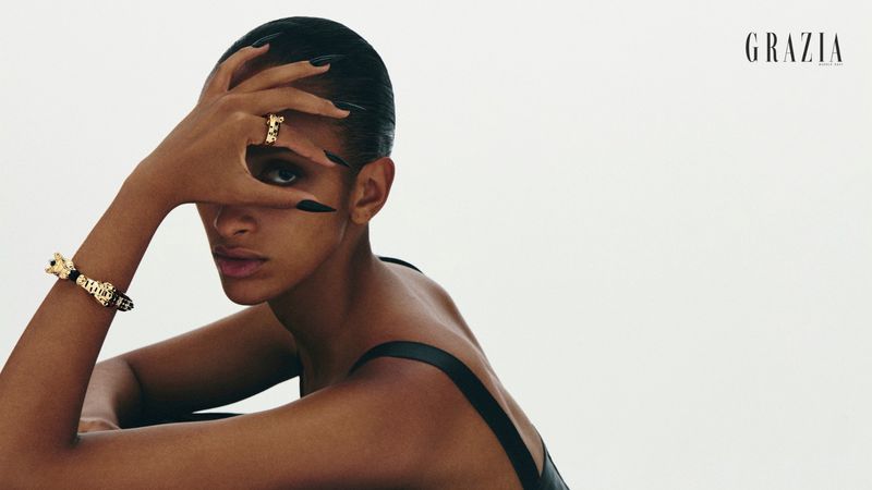 Model with long dark fingernails partially covering face, wearing gold rings and bracelets against a white background