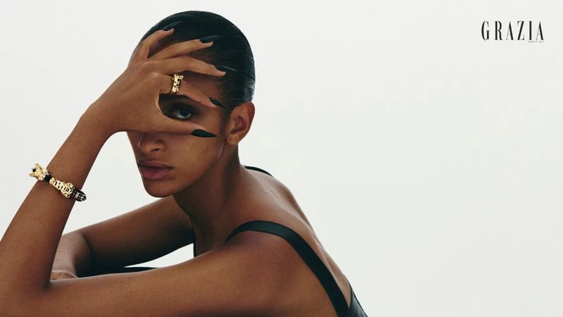 Model with long dark fingernails partially covering face, wearing gold rings and bracelets against a white background