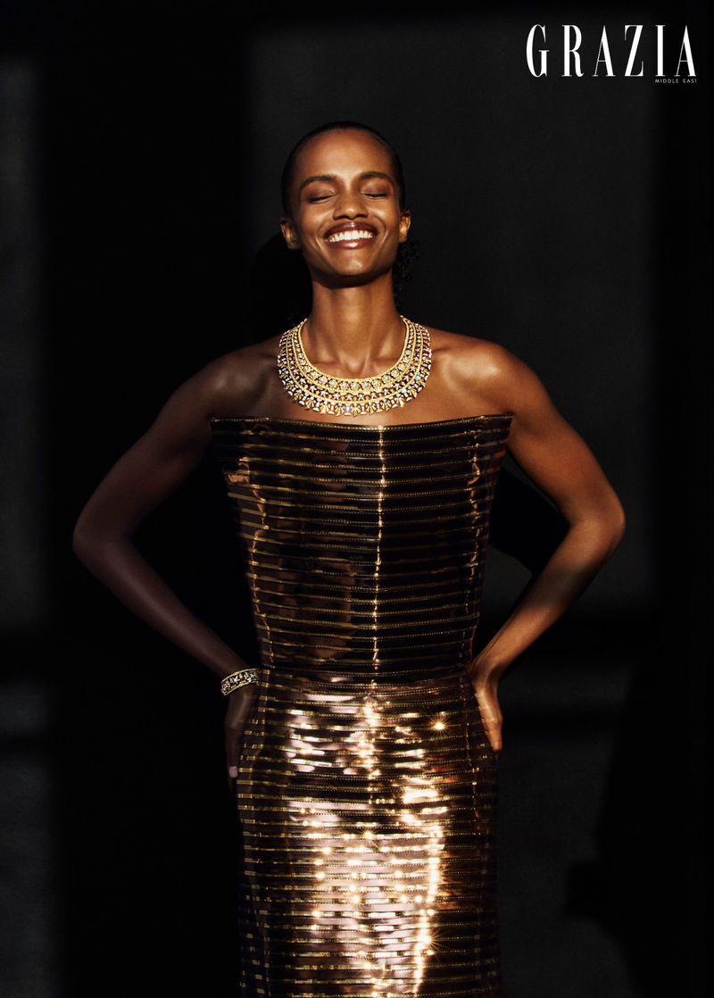 Model smiling with eyes closed, wearing a metallic gold strapless dress and statement necklace against a dark background