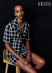 Model Farhiya Shire seated on a wooden stool wearing a black and white checked tweed jacket and matching shorts against a dark background.