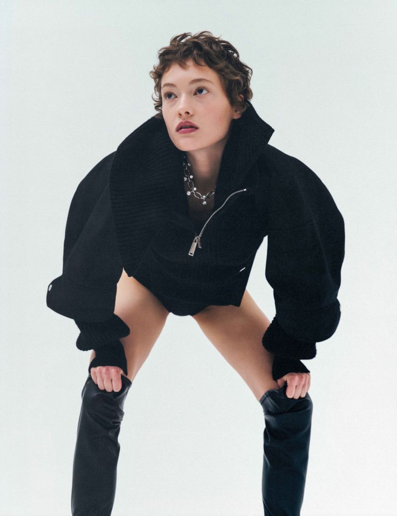 Model with short curly brown hair wearing a black jacket and leather boots against a white background