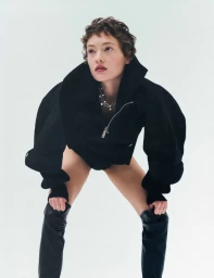 Model with short curly brown hair wearing a black jacket and leather boots against a white background