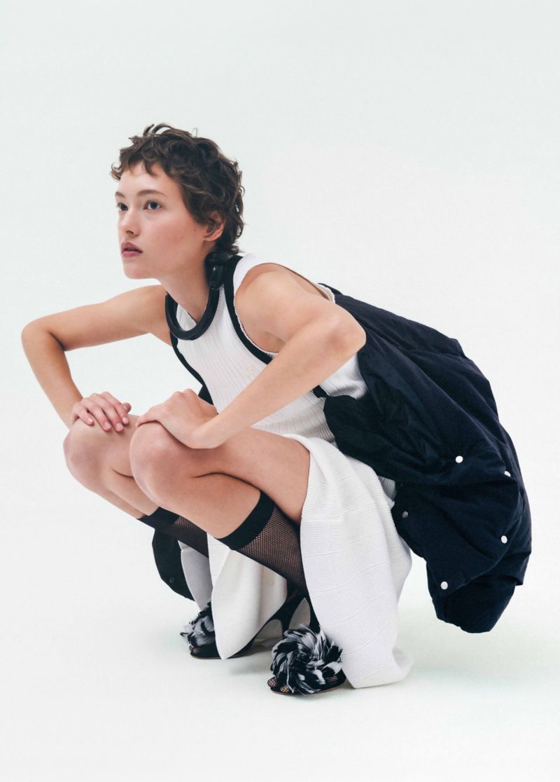 Model with short dark hair wearing a white dress and black jacket squatting against a plain white studio backdrop.