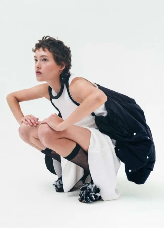 Model with short dark hair wearing a white dress and black jacket squatting against a plain white studio backdrop.