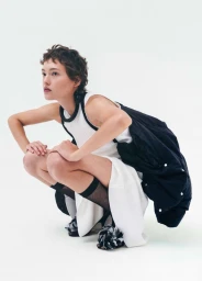 Model with short dark hair wearing a white dress and black jacket squatting against a plain white studio backdrop.