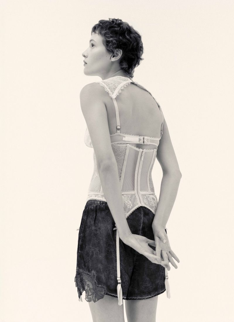 Model with short dark hair wearing a lace-trimmed corset top and dark shorts against a pale background
