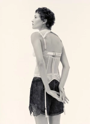 Model with short dark hair wearing a lace-trimmed corset top and dark shorts against a pale background