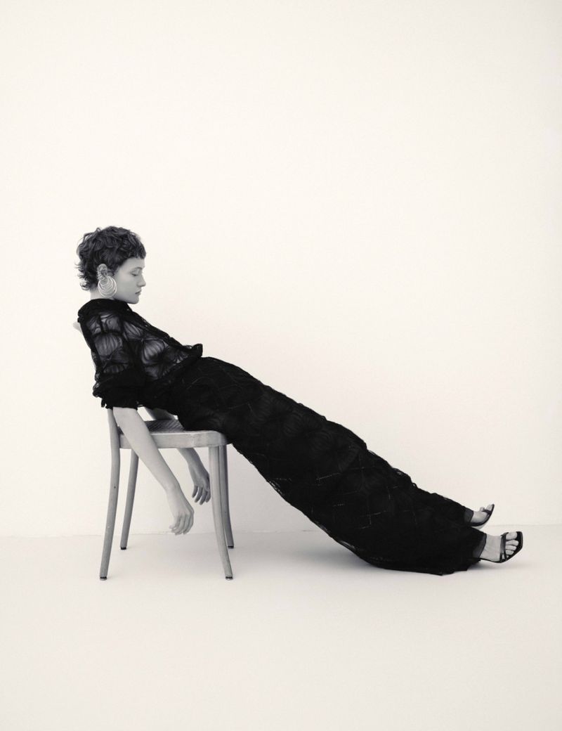 Model with short curly hair wearing a long black dress reclining backwards on a wooden chair against a plain white wall