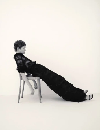 Model with short curly hair wearing a long black dress reclining backwards on a wooden chair against a plain white wall